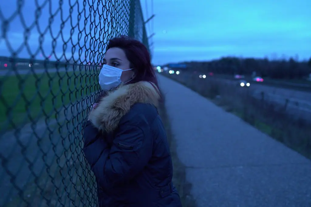 image of a woman wearing a surgical mask leaning on a chainlink fence looking longingly into the distance